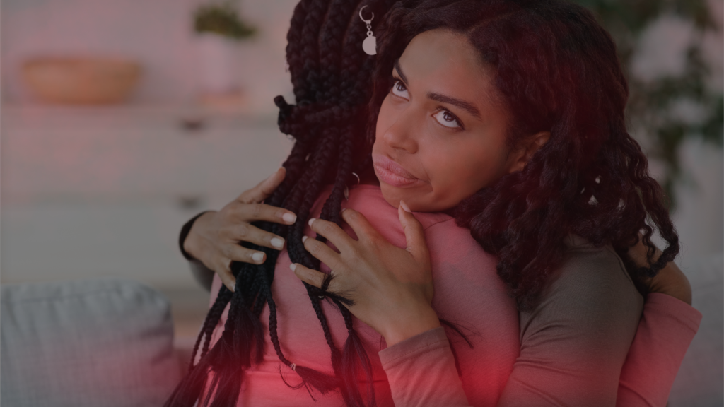 Duas mulheres se abraçam, mas uma delas exibe uma expressão de desconfiança e insatisfação, sugerindo falsidade ou intenções ocultas. A cena representa a inveja disfarçada de amizade e sua influência negativa nos relacionamentos.