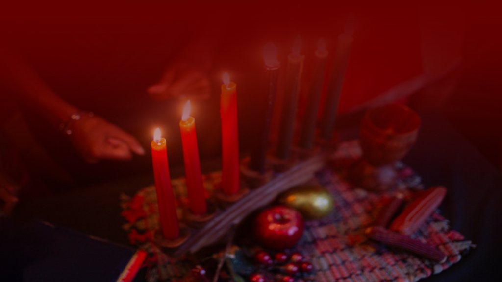 Imagem de um altar com velas vermelhas acesas em destaque, dispostas ao lado de frutas, um cálice de madeira e elementos decorativos tradicionais. As mãos desfocadas no fundo sugerem um ritual sendo realizado, enquanto os tons de vermelho criam uma atmosfera de intensidade e conexão espiritual.