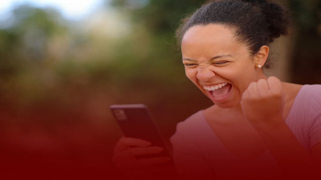 Mulher sorridente olhando para seu celular com expressão de euforia e felicidade, comemorando enquanto levanta o punho em sinal de vitória. O fundo desfocado sugere um ambiente externo, enquanto os tons de vermelho na parte inferior reforçam a intensidade emocional da cena.