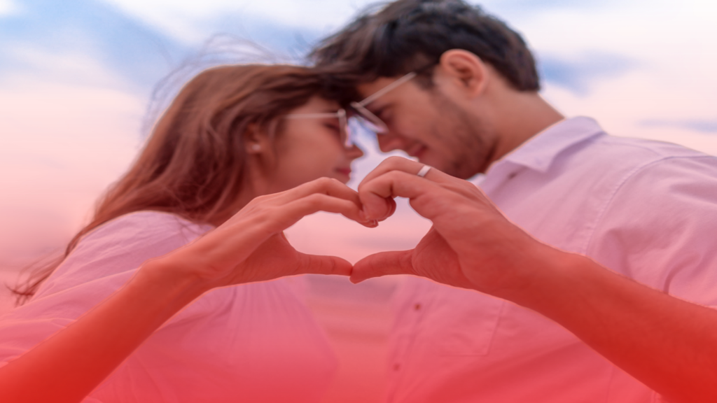 Um casal em um momento romântico, formando um coração com as mãos em primeiro plano. Eles se olham carinhosamente, com um fundo de céu suave e tons de rosa e vermelho que evocam a ideia de o Selamento Espiritual é o único ritual que tem como trazer o ex de volta de forma definitiva. 