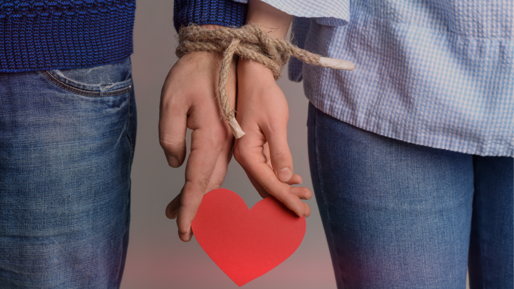 Duas mãos entrelaçadas segurando um coração vermelho de papel, enquanto estão amarradas juntas por uma corda rústica. O casal veste roupas casuais, com jeans e blusas de manga longa, transmitindo uma sensação de vínculo e conexão. O fundo neutro destaca a simbologia do amor e do compromisso.