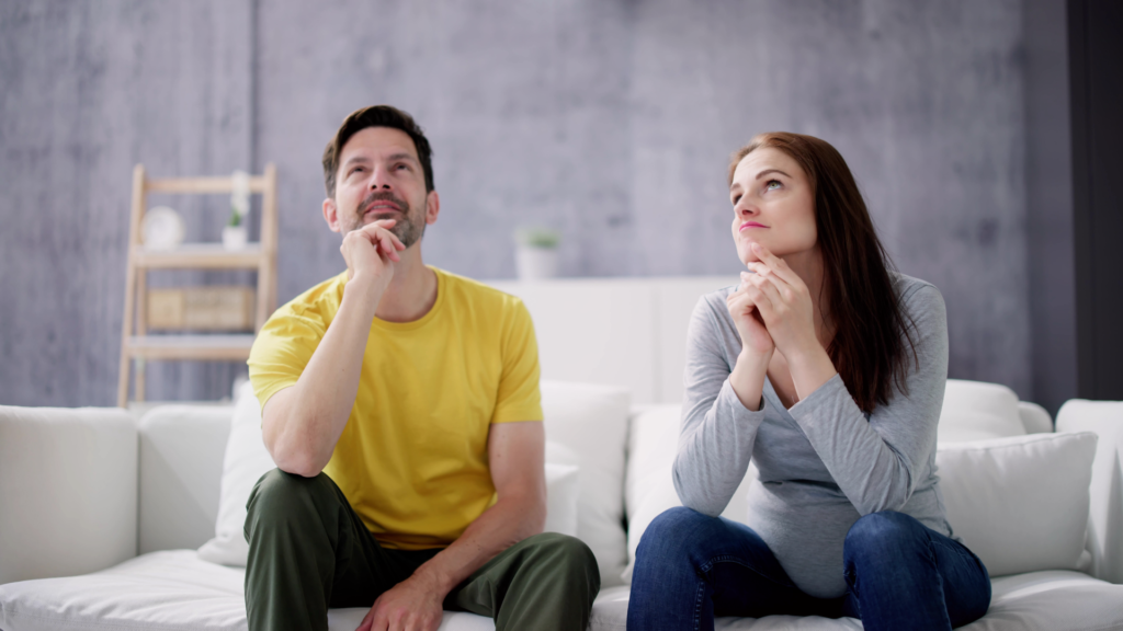 Um homem e uma mulher sentados em um sofá, ambos com expressões pensativas, olhando para cima como se estivessem refletindo sobre suas vidas ou experiências passadas.