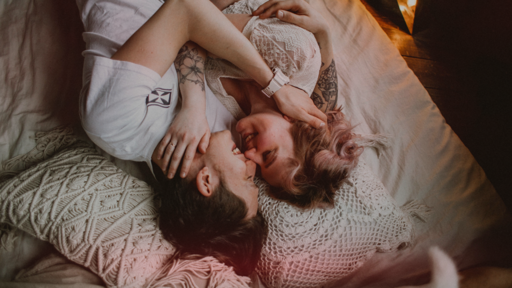 Casal deitado junto na cama, sorrindo e olhando um para o outro com proximidade e carinho. A cena transmite amor, conexão e felicidade, sugerindo que existe um antídoto para relacionamento que esfriou.