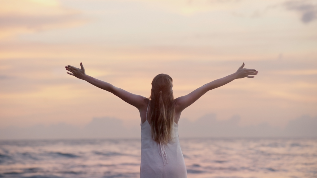 Uma mulher de costas, vestindo um vestido branco, abre os braços em direção ao horizonte do mar ao pôr do sol. A imagem transmite liberdade, renovação e um novo começo, simbolizando a superação e a escolha de um futuro melhor.