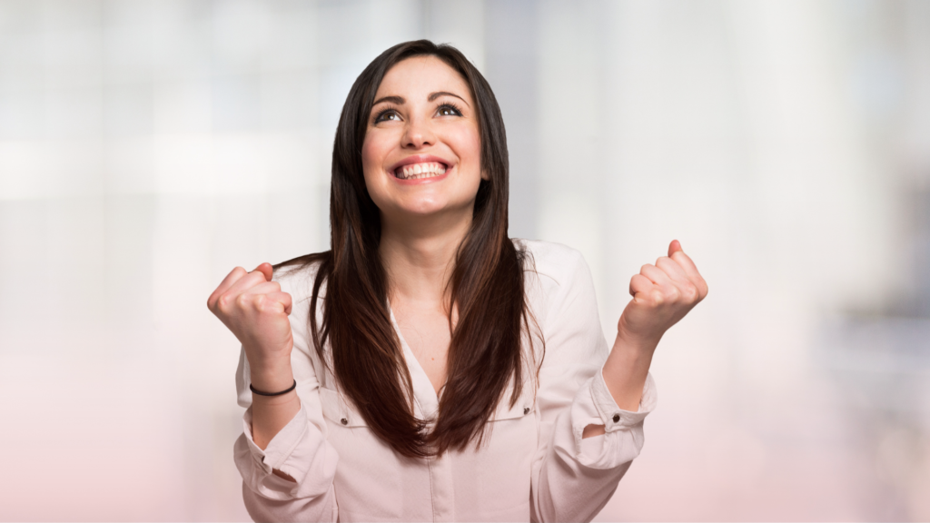 Mulher sorridente com expressão de felicidade e empolgação, olhando para cima com os punhos cerrados em um gesto de celebração.