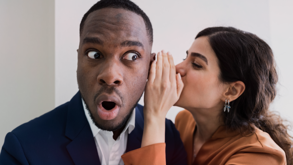 Homem com expressão surpresa e desconfiada enquanto uma mulher sussurra algo em seu ouvido. Ele veste um terno azul e uma camisa branca, enquanto a mulher, que está próxima a ele, veste um blazer marrom e brincos prateados. O homem olha para o lado, sugerindo surpresa ou dúvida sobre o que está ouvindo. A cena transmite a ideia de fofoca, manipulação ou tentativa de criar desconfiança.
