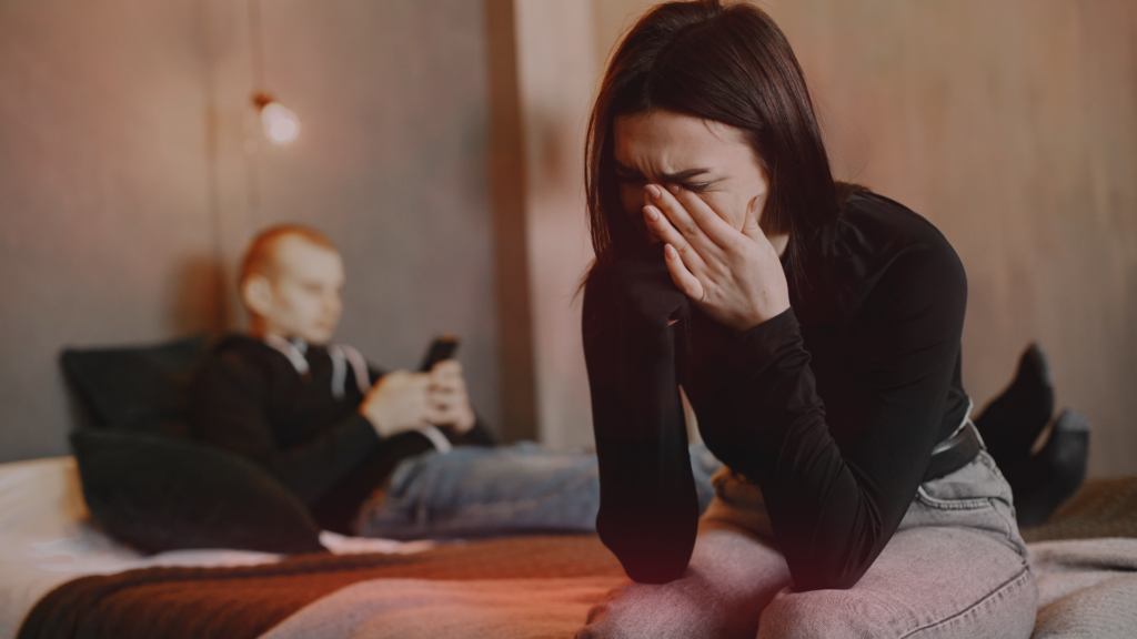 Mulher sentada na beira da cama, com o rosto coberto pelas mãos, expressando tristeza e frustração. Ao fundo, um homem deitado, distraído com o celular, ignorando sua presença. A cena transmite afastamento emocional e falta de sexo no relacionamento.