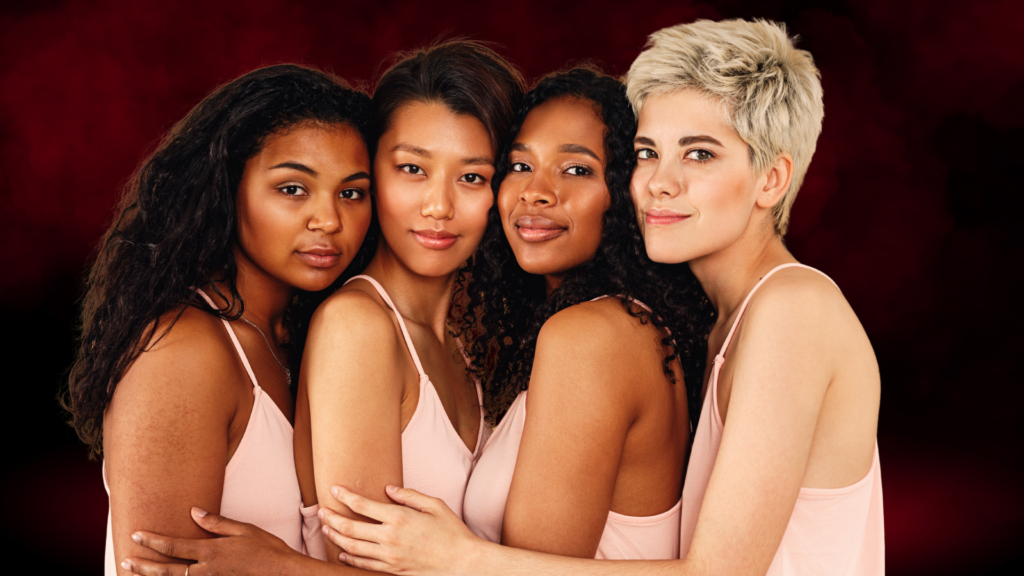 Quatro mulheres de diferentes origens e estilos se abraçam, transmitindo união, confiança e empoderamento. Todas vestem roupas claras, contrastando com o fundo em tons de vermelho escuro e preto.