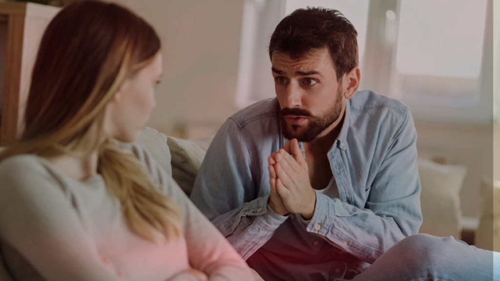 Um homem de expressão arrependida, com as mãos unidas em um gesto de súplica, olha para uma mulher sentada ao seu lado. Ela mantém os braços cruzados e seu rosto expressa dúvida e distanciamento, refletindo um dilema emocional, se deve perdoar a traição. O ambiente é intimista e acolhedor, mas a tensão entre o casal é evidente.