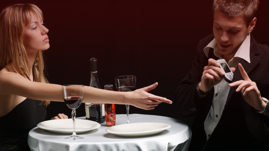 Um casal jantando em um ambiente sofisticado, com uma garrafa de vinho sobre a mesa. A mulher, com expressão séria e olhar incisivo, estende a mão em direção ao homem, que segura um celular e faz um gesto de defesa com a outra mão, como se estivesse tentando se justificar. O clima da cena sugere tensão, desconfiança e possível conflito causado pelo ciúme.

