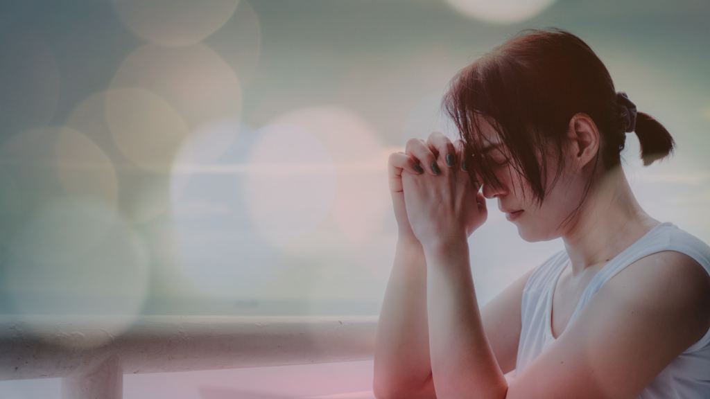 Mulher de olhos fechados e mãos unidas em uma posição de reflexão ou oração, em um ambiente calmo e iluminado com efeitos de luz suave ao fundo, evocando um momento de espiritualidade e introspecção.