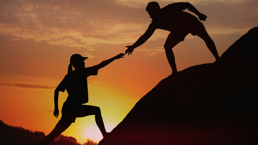 Silhueta de duas pessoas em uma encosta durante o pôr do sol, onde uma delas está estendendo a mão para ajudar a outra a subir. A cena é marcada por tons quentes e dourados do entardecer, simbolizando apoio, parceria e superação em momentos difíceis.