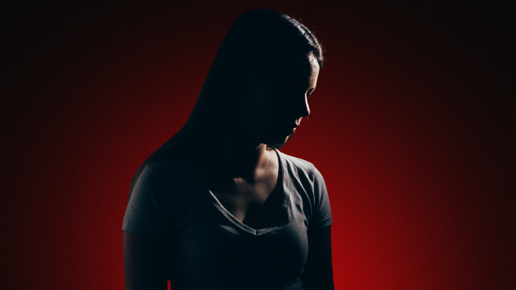 Imagem de uma mulher com o rosto parcialmente coberto por sombras, expressando tristeza e introspecção. O fundo escuro com tons avermelhados cria uma atmosfera emocional intensa, simbolizando dor interna e cicatrizes emocionais não resolvidas.