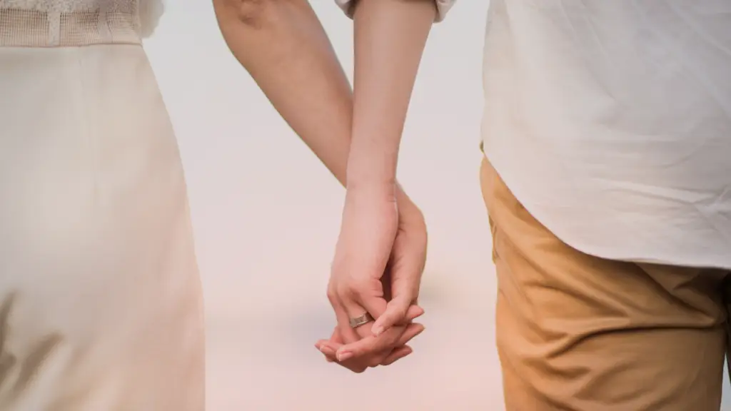 Duas pessoas de mãos dadas, com os dedos entrelaçados, simbolizando carinho, conexão e compromisso. Uma das mãos usa uma aliança prateada, reforçando a ideia de união e amor verdadeiro, depois de perceber os sinais de amor. O fundo suave e claro cria uma atmosfera tranquila e romântica.