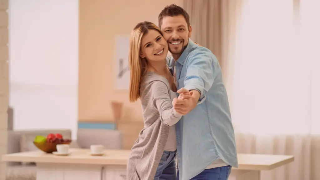 Casal sorridente dançando alegremente dentro de casa, transmitindo leveza, felicidade e conexão saudável. A imagem simboliza liberdade emocional e bem-estar após superar a compulsão emocional, destacando a importância do equilíbrio e da individualidade nos relacionamentos. 