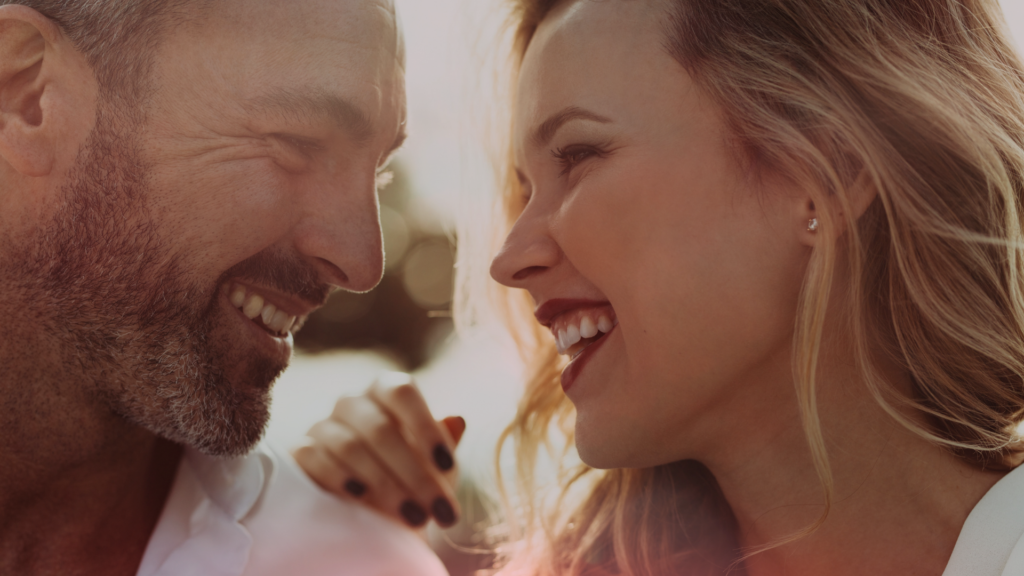 Um casal sorrindo e trocando olhares intensos, demonstrando uma forte conexão emocional. O homem, com barba grisalha e vestindo uma camisa clara, sorri carinhosamente enquanto a mulher, com cabelos loiros e sorriso radiante, o encara com ternura. A luz suave do pôr do sol destaca o momento íntimo entre eles.