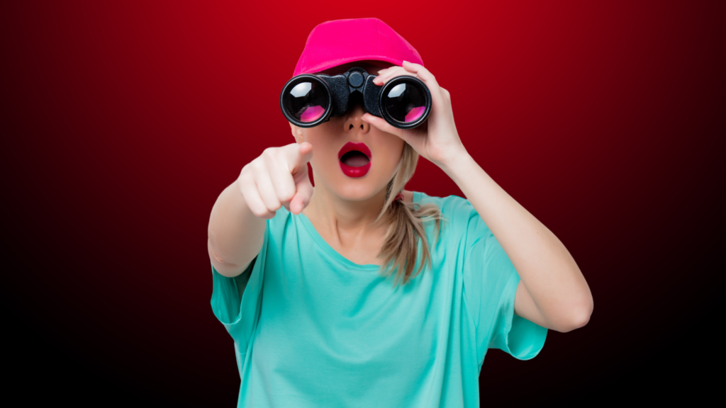 Mulher loira vestindo boné rosa e camiseta verde água, segurando binóculos e apontando para frente com uma expressão surpresa, como que procurando um relacionamento sério. O fundo em tons de vermelho escuro adiciona um ar de intensidade e busca determinada.