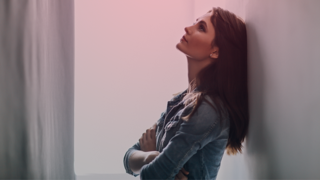 Uma mulher pensativa, encostada na parede, olhando para cima com expressão introspectiva e melancólica. Seu semblante transmite um misto de tristeza e reflexão, sugerindo um momento de angústia emocional ou incerteza amorosa por causa de um relacionamento platônico 