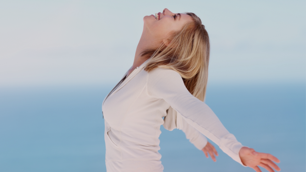 Mulher loira de braços abertos e rosto voltado para o céu, sorrindo com leveza diante de um fundo azul, simbolizando liberdade, reconexão interior e paz após a superação de uma prisão emocional.