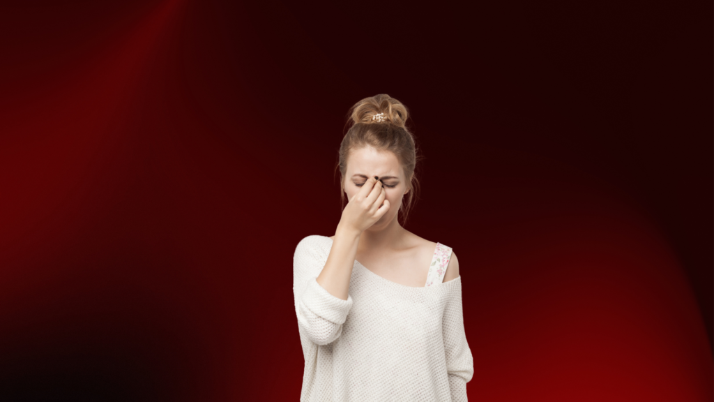 Mulher jovem com expressão de frustração e dor emocional, apertando o topo do nariz com os olhos fechados, transmitindo cansaço, tristeza ou decepção. Usa um suéter branco de ombro caído. O fundo vermelho escuro reforça a intensidade emocional da cena.