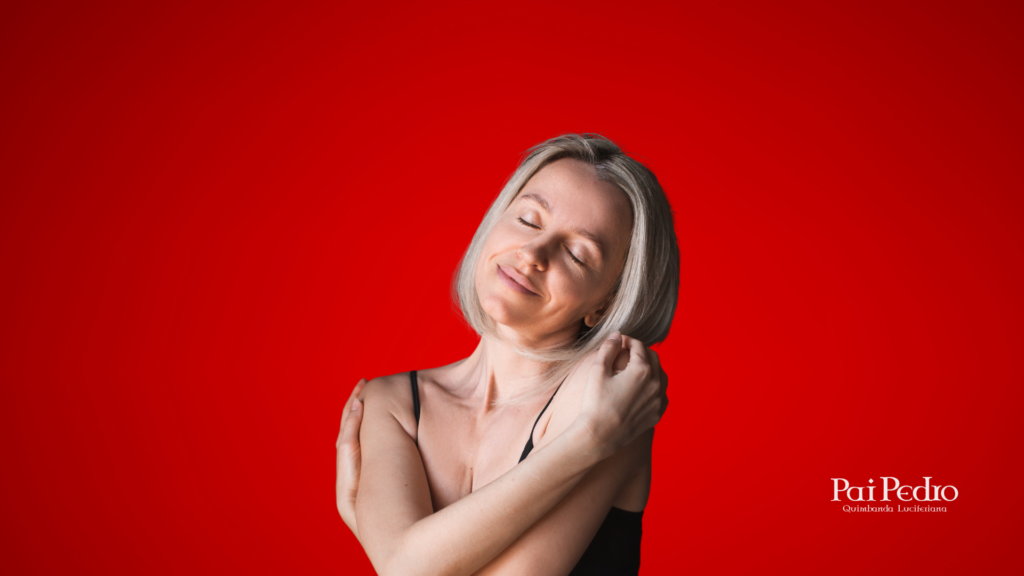 Mulher de cabelos claros sorrindo suavemente, de olhos fechados, abraçando a si mesma diante de um fundo vermelho intenso. A imagem transmite amor-próprio, paz interior e força emocional.