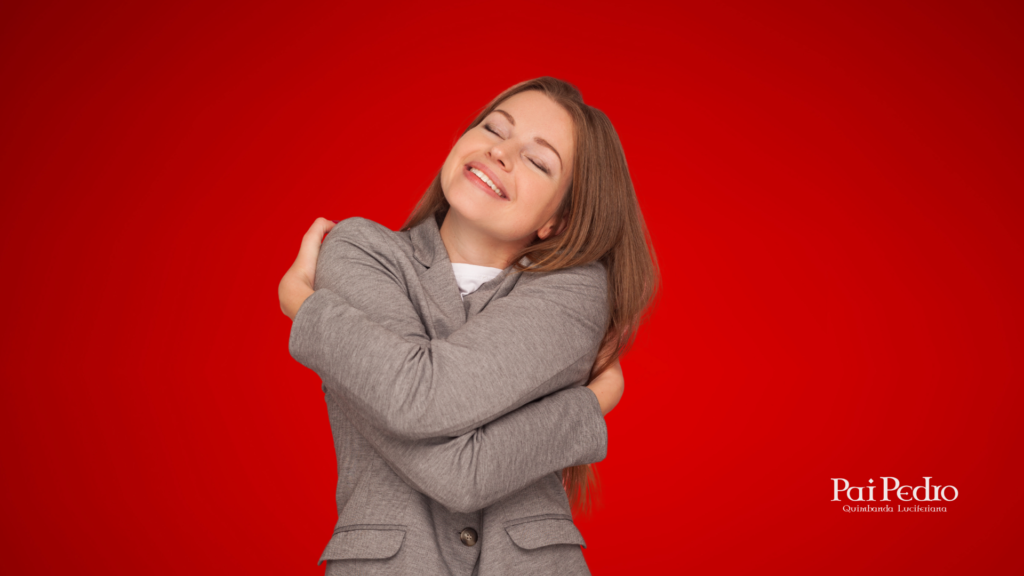 Mulher sorridente de olhos fechados, abraçando a si mesma com ternura diante de um fundo vermelho. A imagem expressa autoestima, acolhimento e a integração  da vibração das mulheres.