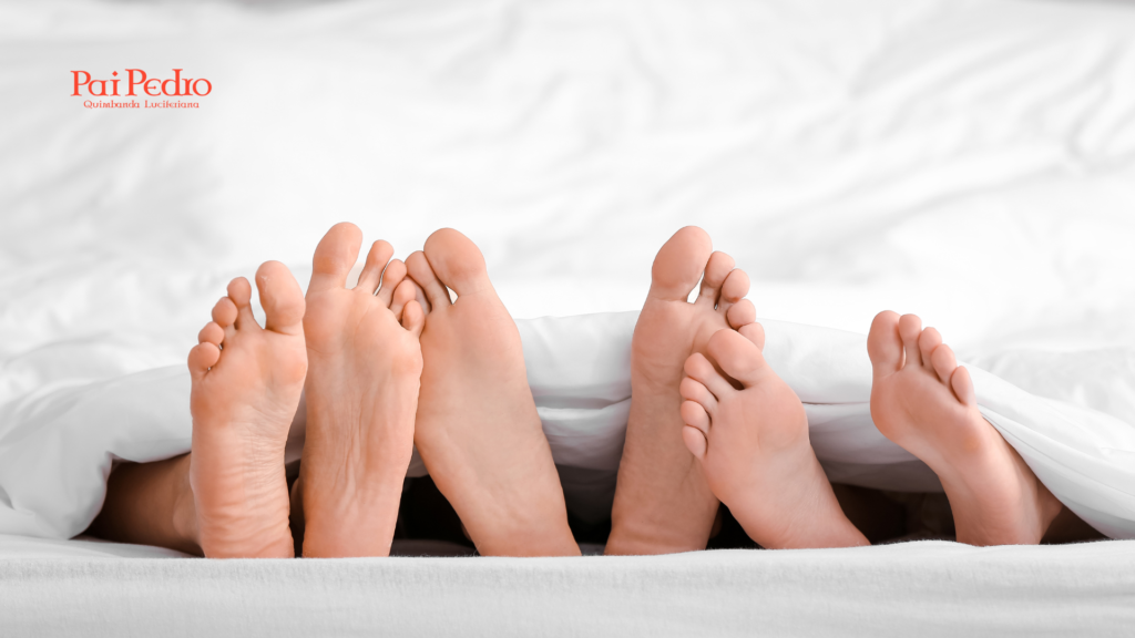 Quatro pés de duas pessoas diferentes sob o mesmo lençol branco, simbolizando intimidade compartilhada, liberdade, mas também possíveis dilemas emocionais de um relacionamento aberto.