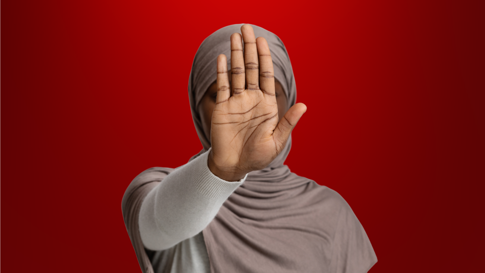 Mulher com expressão séria e mão contínua em sinal de pare, usa um hijab cinza claro, com fundo vermelho intenso, simbolizando alerta e proteção contra um falso pai de santo.