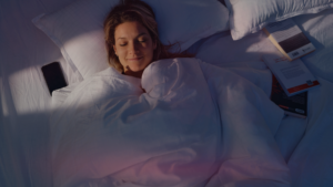 Uma mulher está dormindo serenamente em uma cama iluminada pela luz do entardecer, com um celular ao lado dela no colchão e livros espalhados ao redor. O ambiente transmite uma sensação alegra de sonhar com o ex