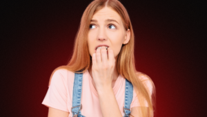 Imagem de uma jovem mulher com expressão ansiosa e preocupada, mordendo as unhas e olhando para o lado. O fundo escuro com tons avermelhados reforça uma atmosfera de tensão e insegurança no relacionamento