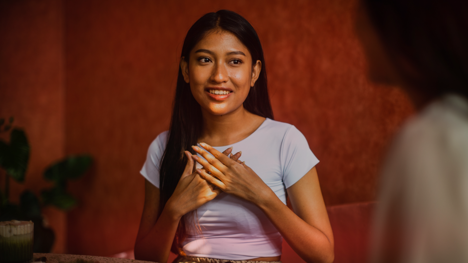 Uma jovem sorridente, com cabelos longos e escuros, usando uma blusa clara, com as mãos sobre o peito em um gesto que expressa emoção e sinceridade, percebendo os sinais de amor. Ela está sentada em um ambiente aconchegante, com uma parede de tom avermelhado ao fundo, conversando com outra pessoa que aparece parcialmente na imagem.