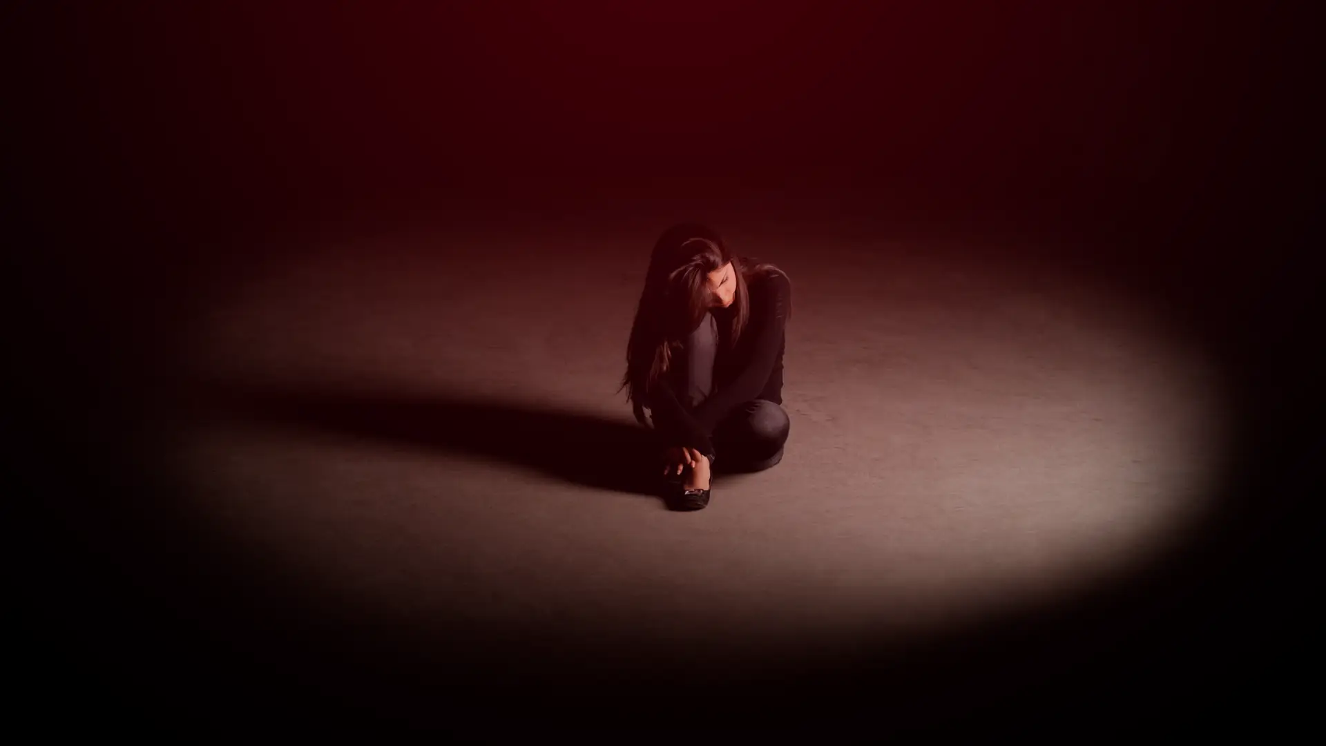 Mulher sentada sozinha no chão, com a cabeça baixa e expressão corporal de tristeza, envolta por uma iluminação escura que destaca seu isolamento emocional.