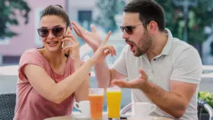 Homem e mulher sentados em uma cafeteria ao ar livre. A mulher, sorridente e despreocupada, fala ao celular enquanto o homem ao lado demonstra irritação e frustração, gesticulando com as mãos, simbolizando o relacionamento rebote. Ambos usam óculos escuros. Há sucos e xícaras sobre a mesa.