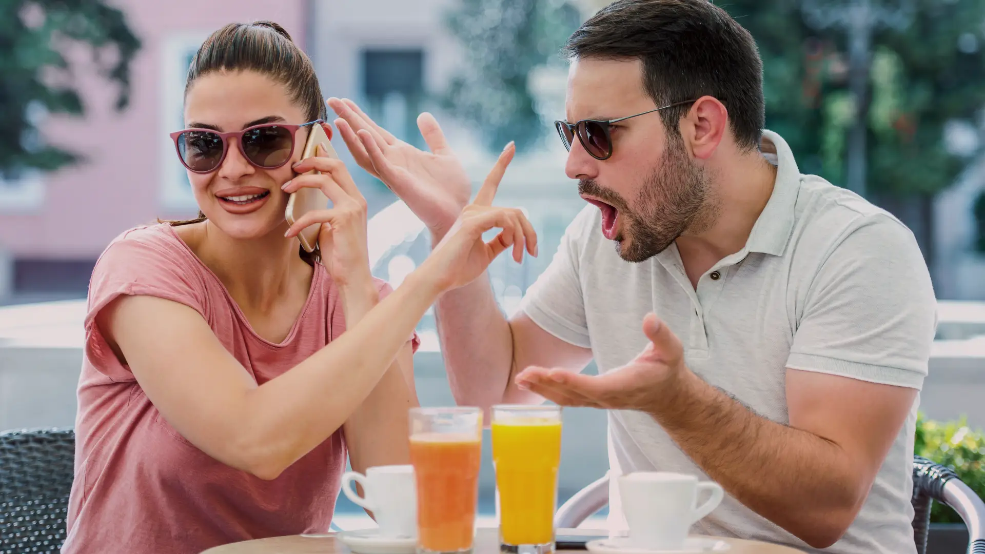 Homem e mulher sentados em uma cafeteria ao ar livre. A mulher, sorridente e despreocupada, fala ao celular enquanto o homem ao lado demonstra irritação e frustração, gesticulando com as mãos, simbolizando o relacionamento rebote. Ambos usam óculos escuros. Há sucos e xícaras sobre a mesa.