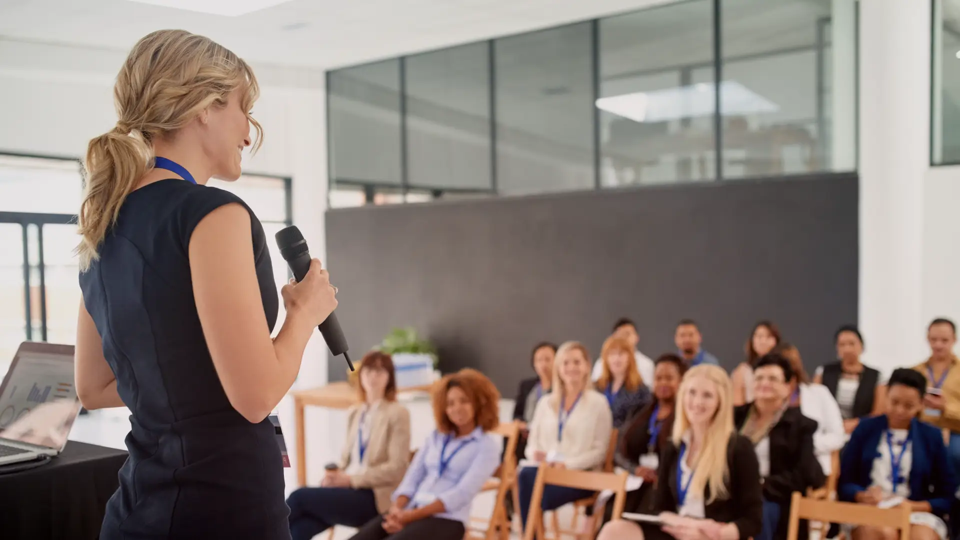 Mulher segurando um microfone e palestrando para um grupo diverso de mulheres atentas em um ambiente corporativo. A cena transmite liderança feminina, empoderamento e troca de conhecimento, resumindo, mulheres inspiradoras.