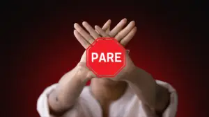 Pessoa com as mãos cruzadas em forma de “X” à frente do rosto, com um sinal de "pare" vermelho sobreposto, simbolizando a decisão firme de contato zero. Fundo em tom escuro com vermelho em destaque, transmitindo urgência e firmeza.