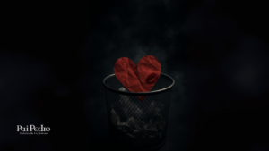 Imagem de um coração vermelho de papel amassado, jogado dentro de uma lixeira metálica. O coração tem a marca de um batom borrado, simbolizando um amor descartado. Fundo escuro e nebuloso, com iluminação dramática destacando apenas o objeto principal, transmitindo de como esquecer alguém que te fez mal.